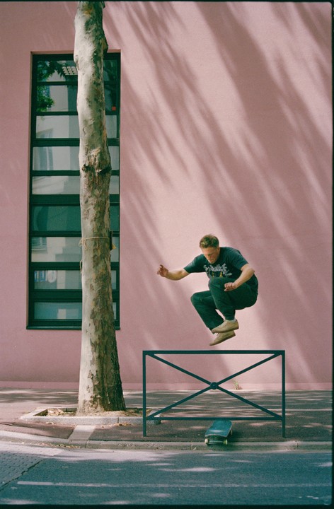 A man jumps over a metal barrier on a street next to a pink building with a tree and a skateboard on the ground.