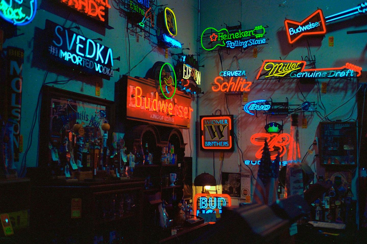 A bar interior is illuminated by numerous neon signs for various alcohol brands.