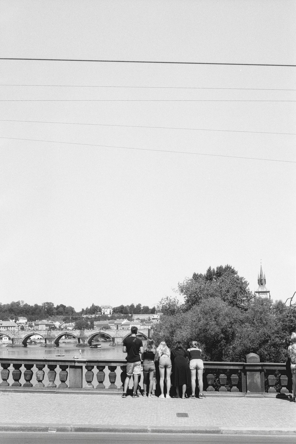 A group of people stand on a bridge overlooking a river with another bridge and a city in the background.