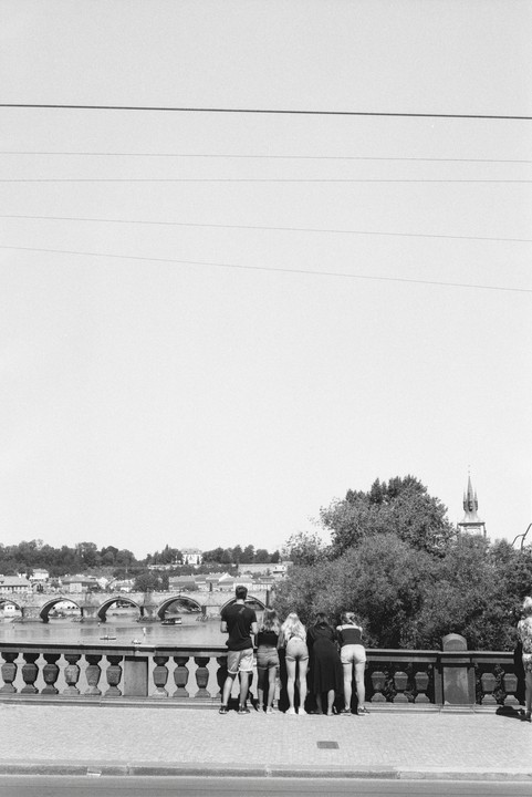 A group of people stand on a bridge overlooking a river with another bridge and a city in the background.