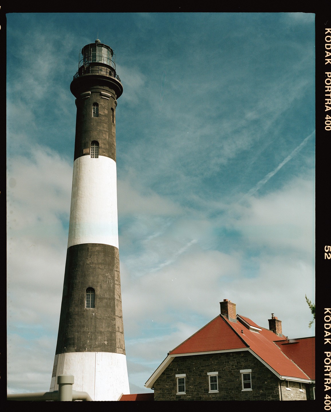 A tall lighthouse with a black and white striped body stands next to a stone building with a red roof under a blue sky.