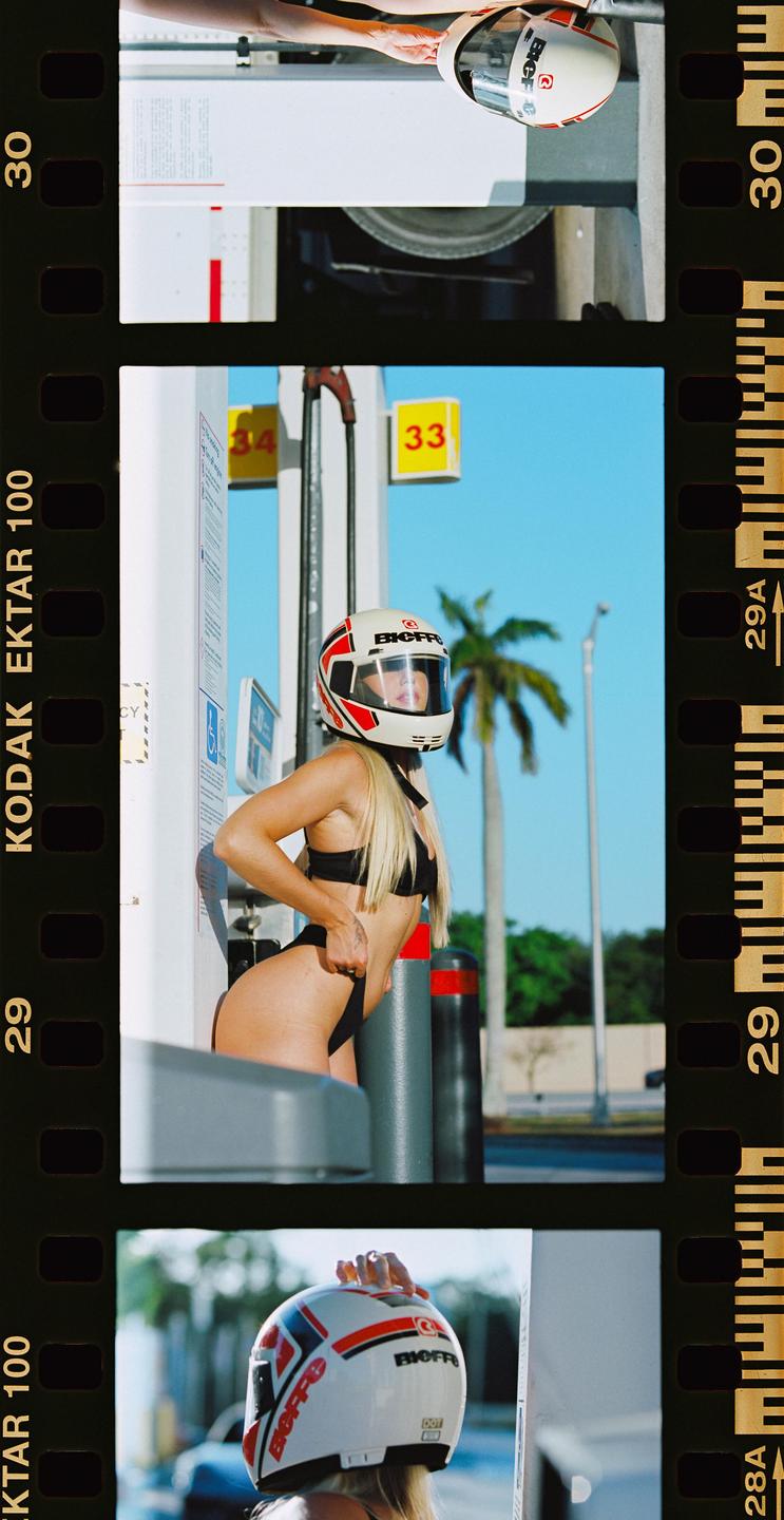 A woman in a bikini and helmet poses at a gas station.