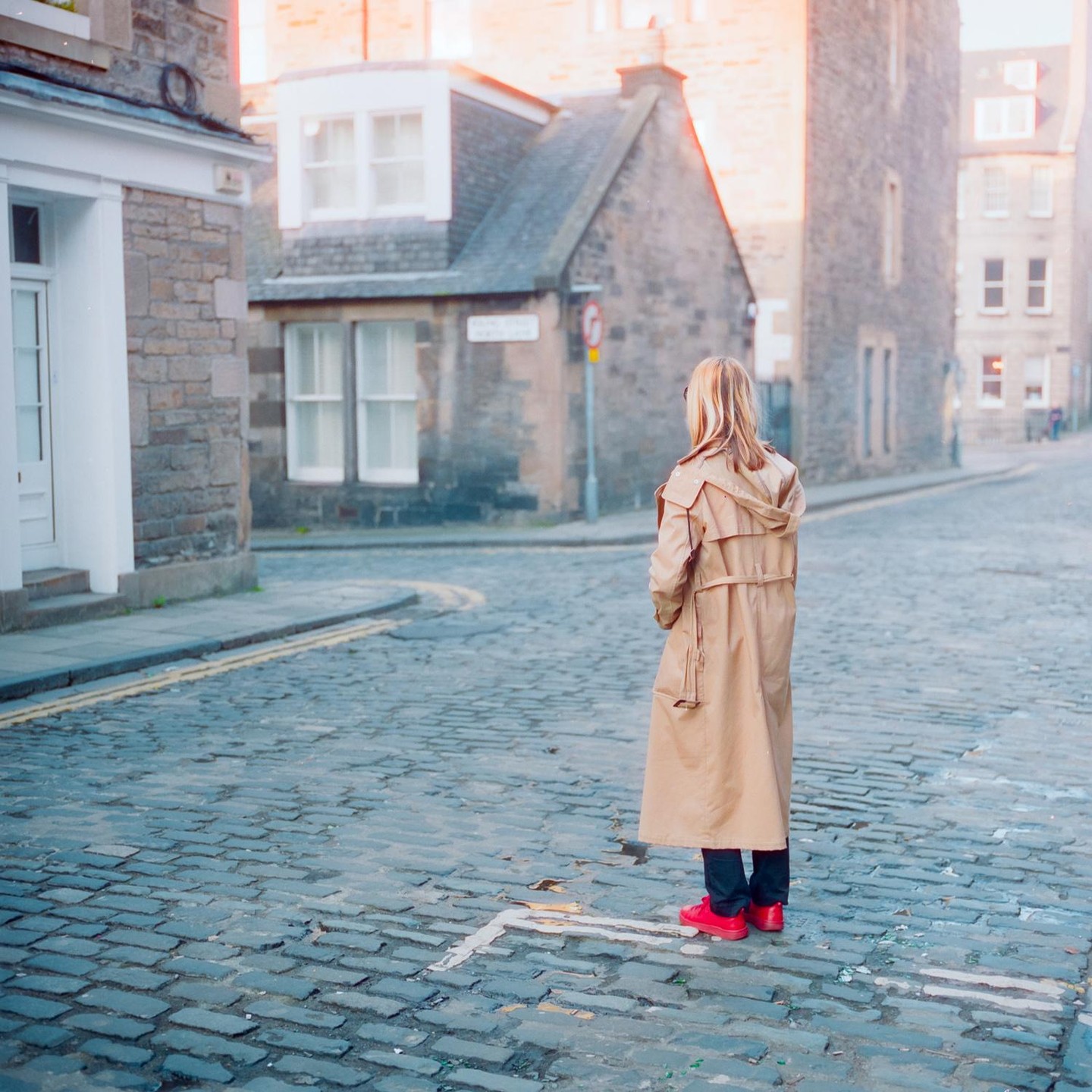 A woman in a trench coat and red shoes stands on a cobblestone street looking towards old buildings.