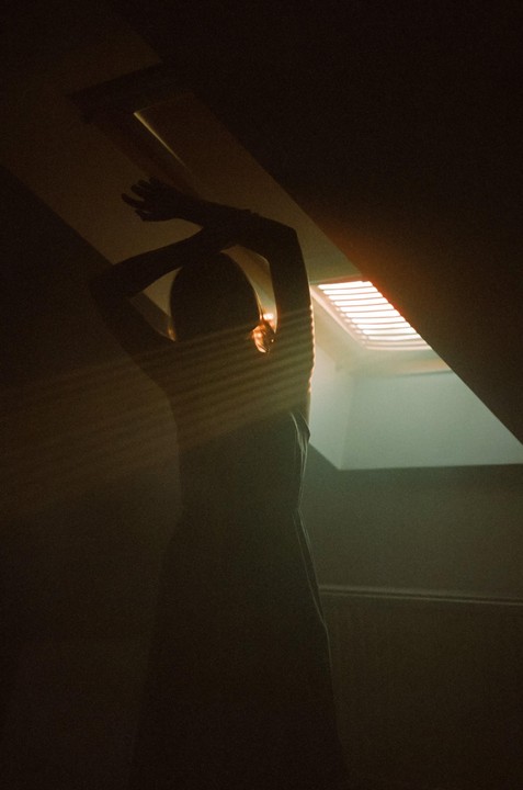 A woman stands in a dark room with her arms raised, illuminated by light rays from a skylight.
