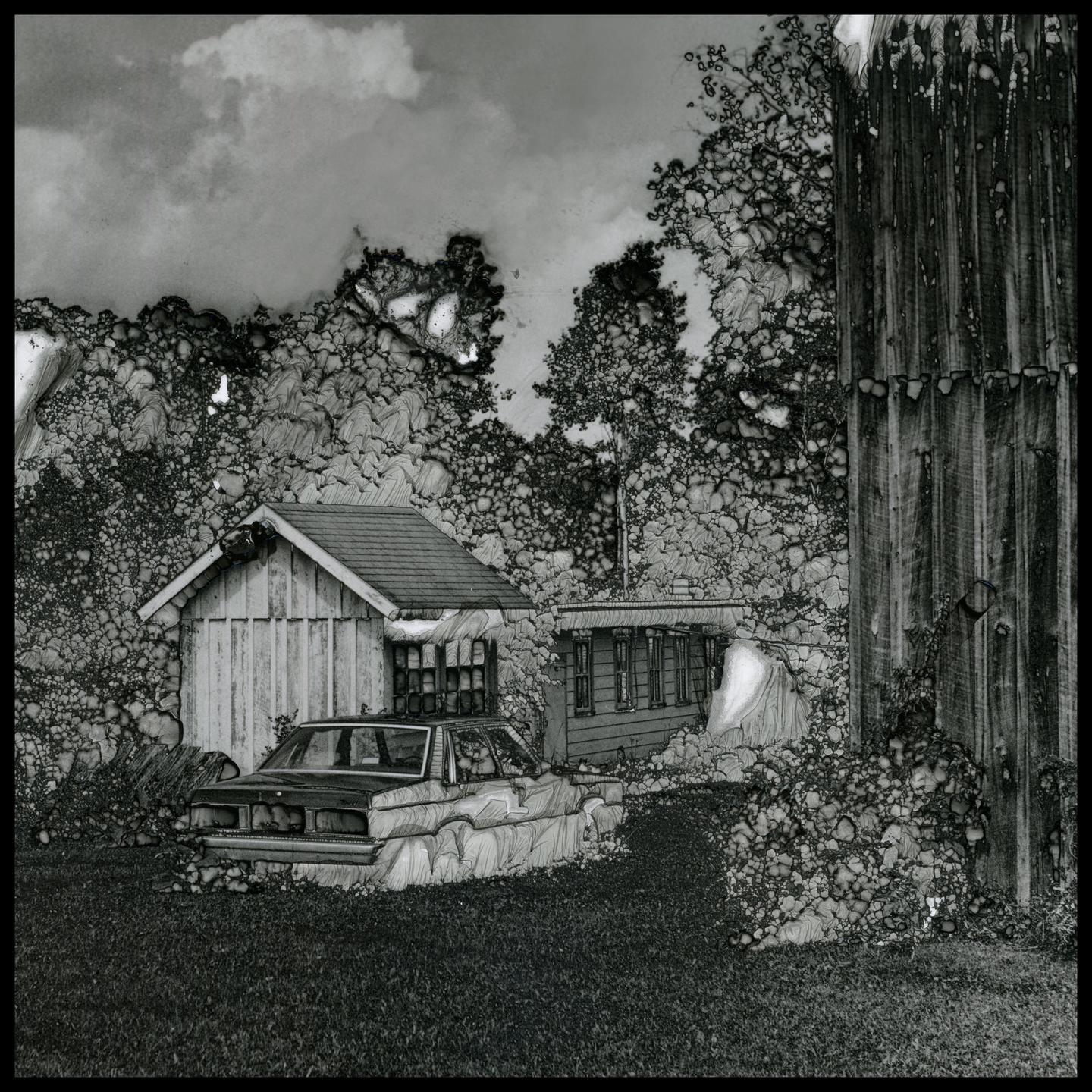 An old car is parked in front of two small buildings with a tall wooden structure on the right.