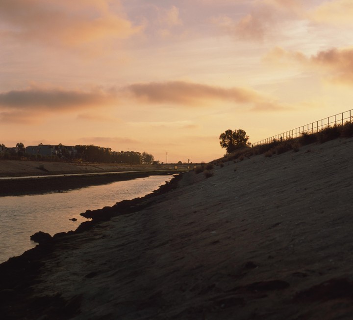 A river flows next to a sandy bank with a fence and a tree under a sunset sky.