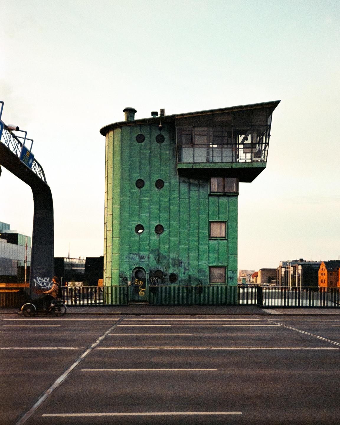 A person rides a bicycle on a bridge next to a tall, green, cylindrical building with a slanted roof and round windows.