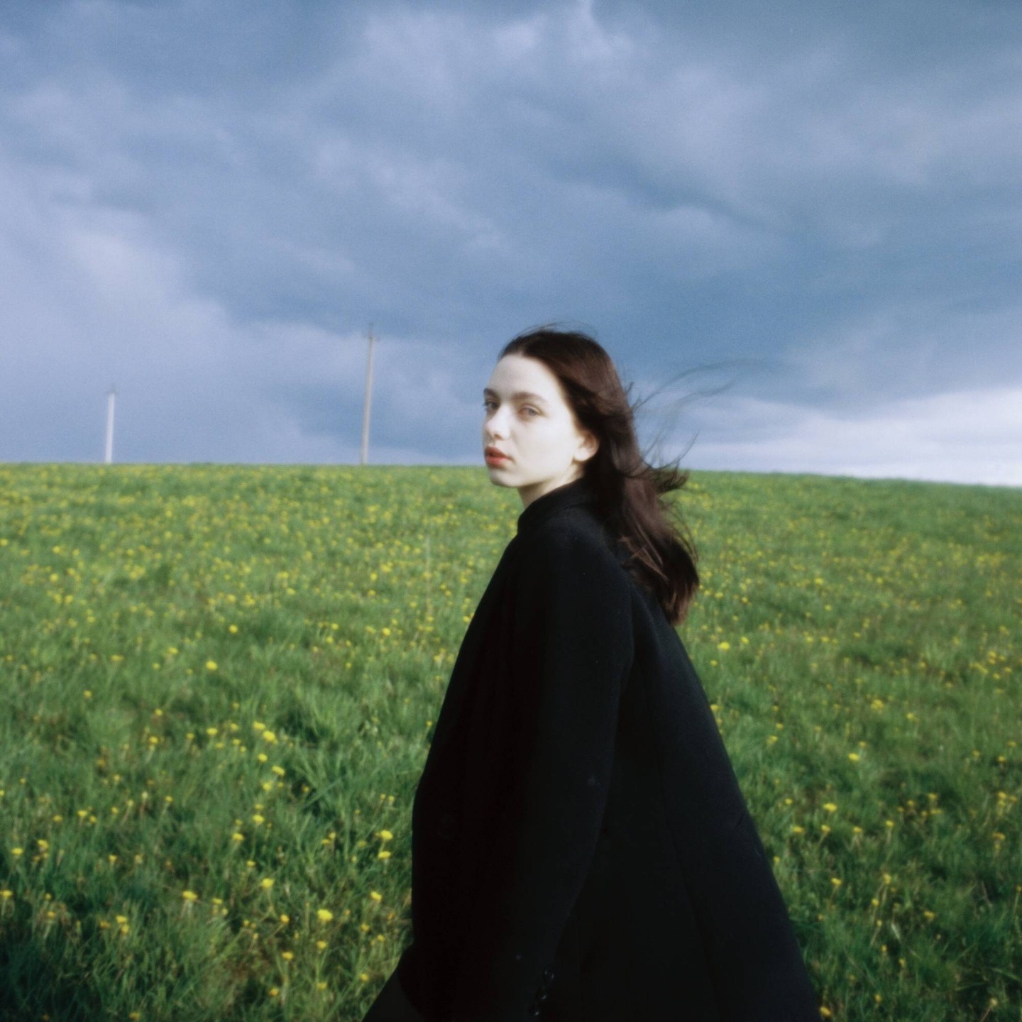 A woman with long hair looks over her shoulder while standing in a field of green grass and yellow flowers under a cloudy sky.