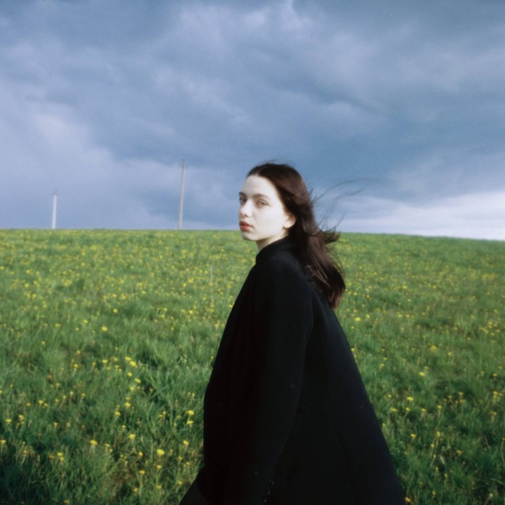 A woman with long hair looks over her shoulder while standing in a field of green grass and yellow flowers under a cloudy sky.