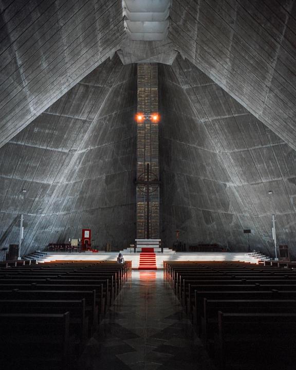 A woman sits in a brutalist cathedral with a red carpet leading to an altar.