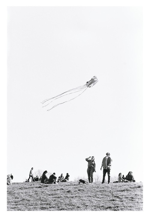 A kite flies high above a grassy hill where people are gathered, some sitting and some standing.