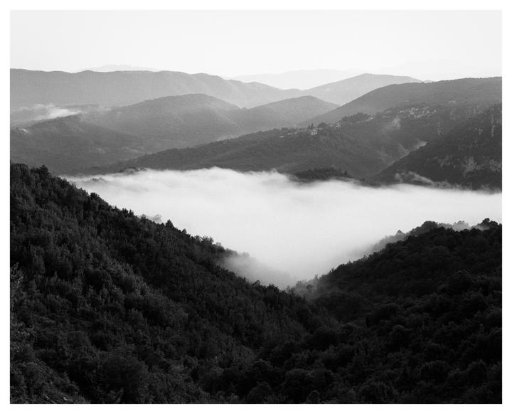 A valley filled with fog is surrounded by forested mountains under a bright sky.