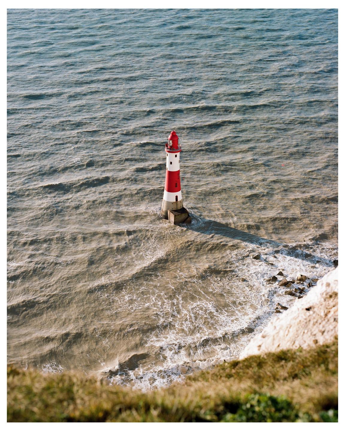 A red and white lighthouse stands in the ocean with waves crashing around its base.