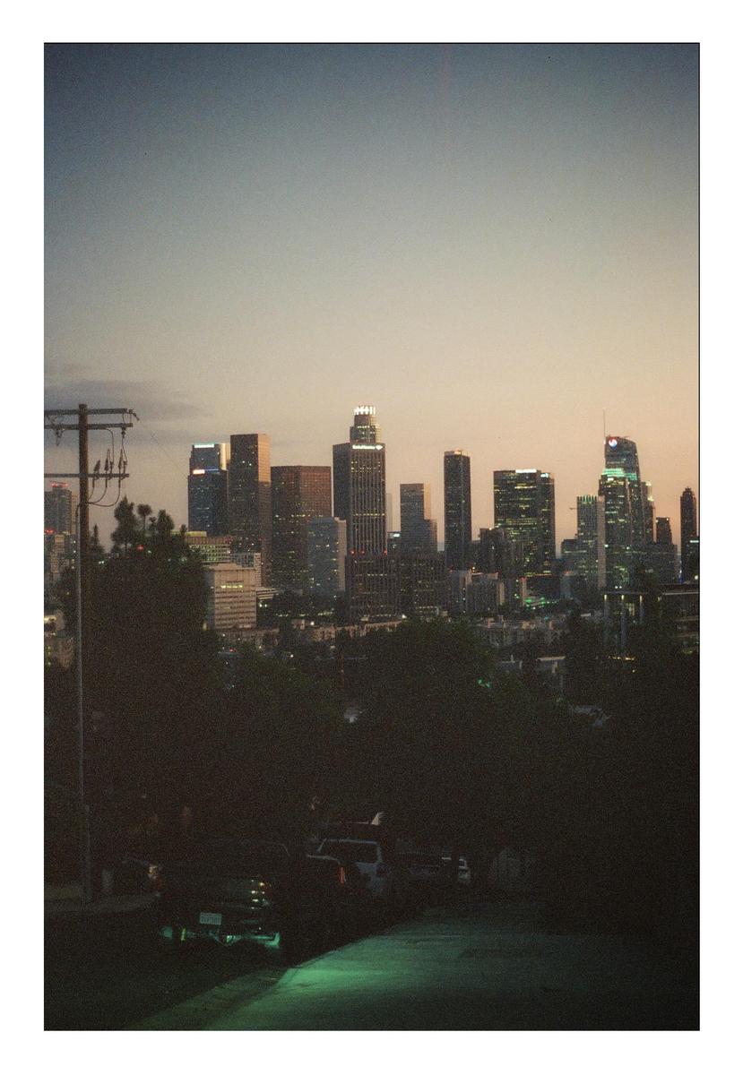 A city skyline with illuminated buildings is visible above a dark street and trees at dusk.