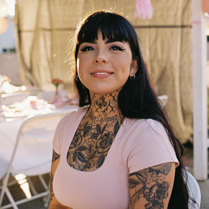 A woman with long dark hair and tattoos on her chest and neck smiles at the camera.