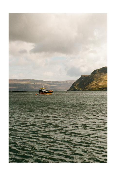 A fishing boat floats on the water with a mountainous coastline in the distance under a cloudy sky.