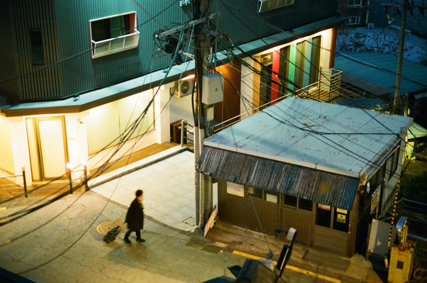 A person walks on a cracked street past a small building and a larger building with colorful windows at night.