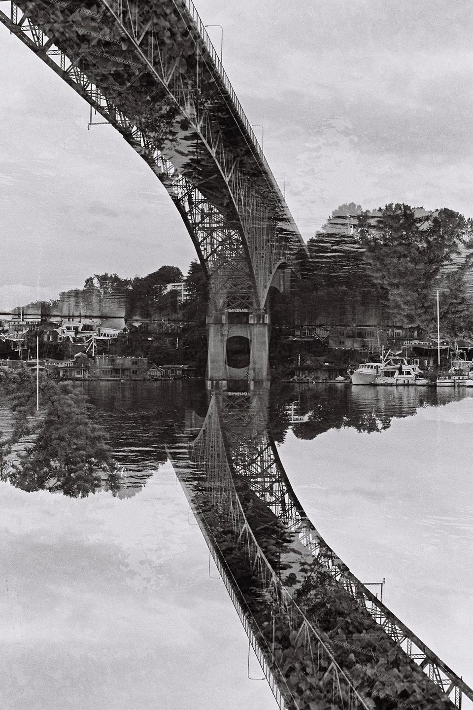 A double exposure shows a bridge over water with boats and houses on the shore, with the bridge reflected in the water.