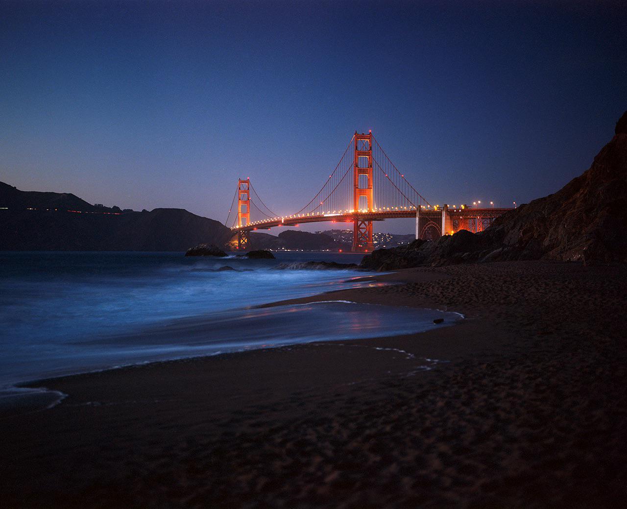 The Golden Gate Bridge is illuminated at night with a beach and ocean in the foreground.