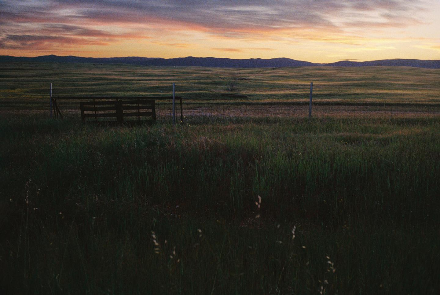 A wooden fence and posts stand in a grassy field with mountains in the distance under a colorful sunset sky.