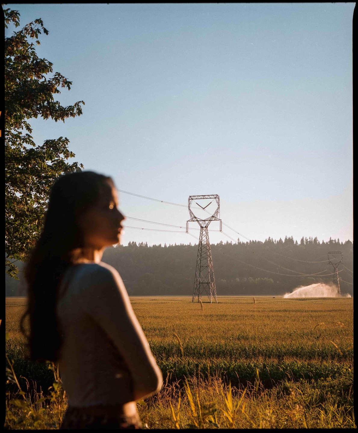 A woman stands in a field with power lines and sprinklers in the background.