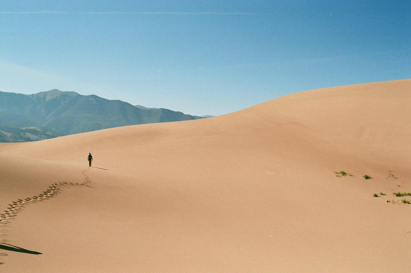 A person walks across a vast sand dune with mountains in the background under a clear sky.