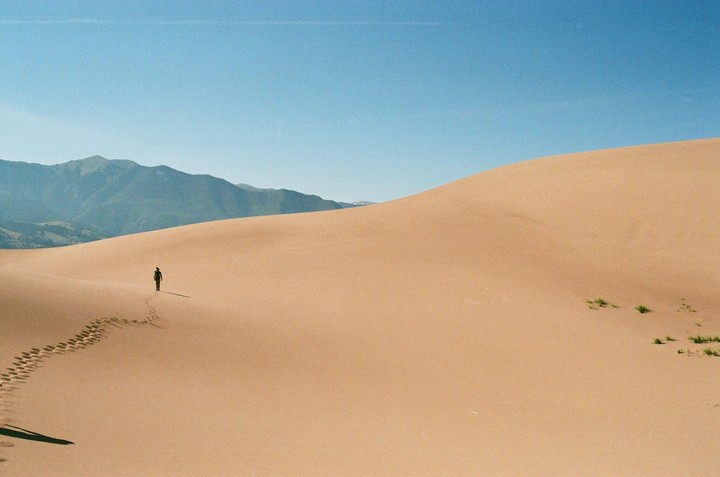 A person walks across a vast sand dune with mountains in the background under a clear sky.