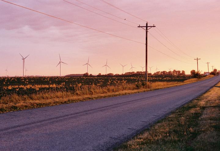 A long road stretches into the distance alongside a field with numerous wind turbines under a colorful sky.