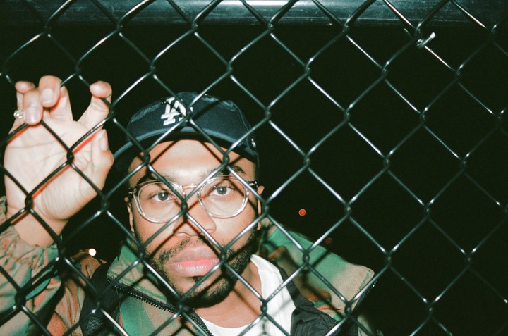 A man wearing glasses and a baseball cap looks through a chain link fence at night.