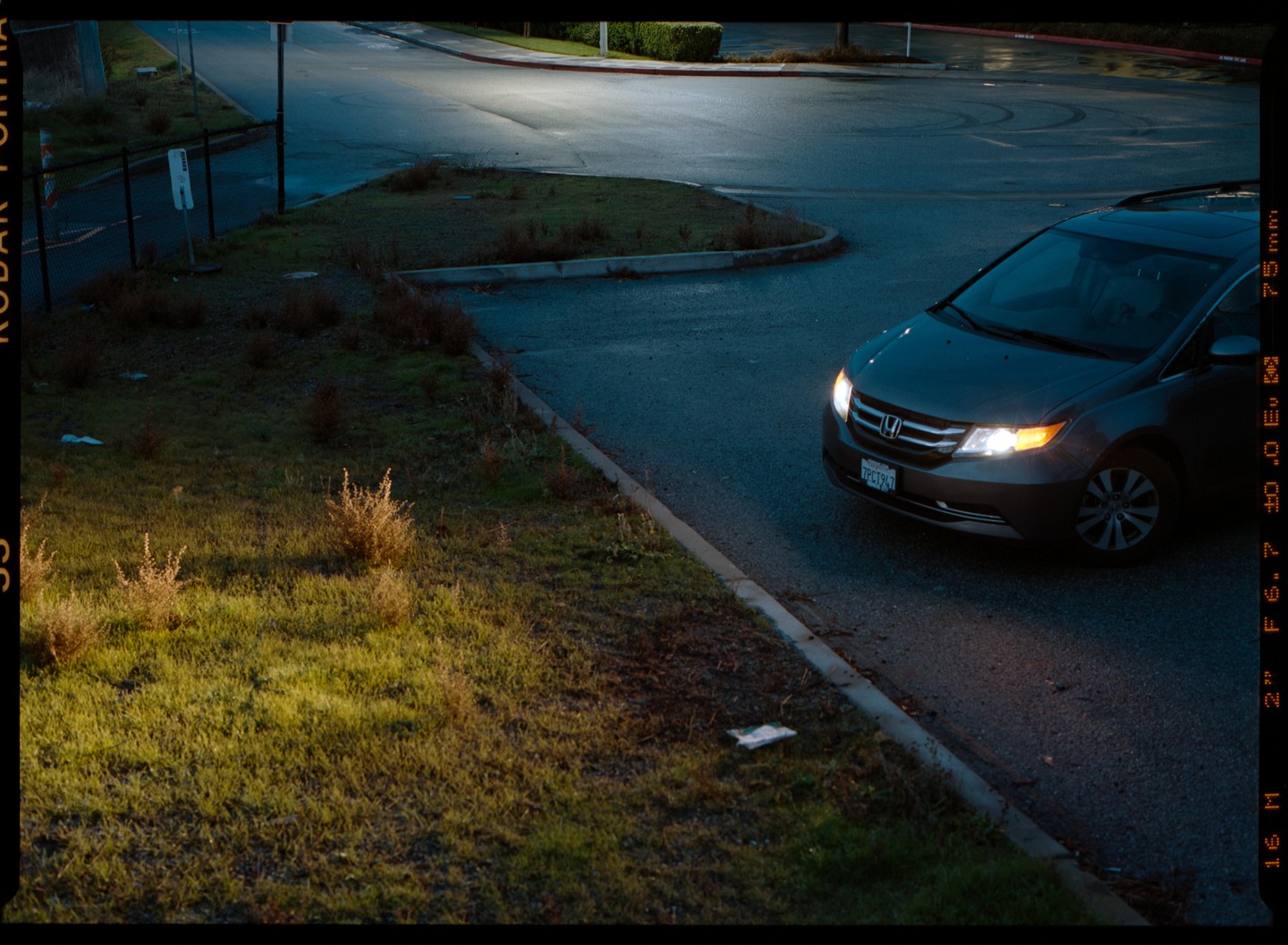 A gray minivan with its headlights on is parked on the side of a road next to a grassy area at dusk.