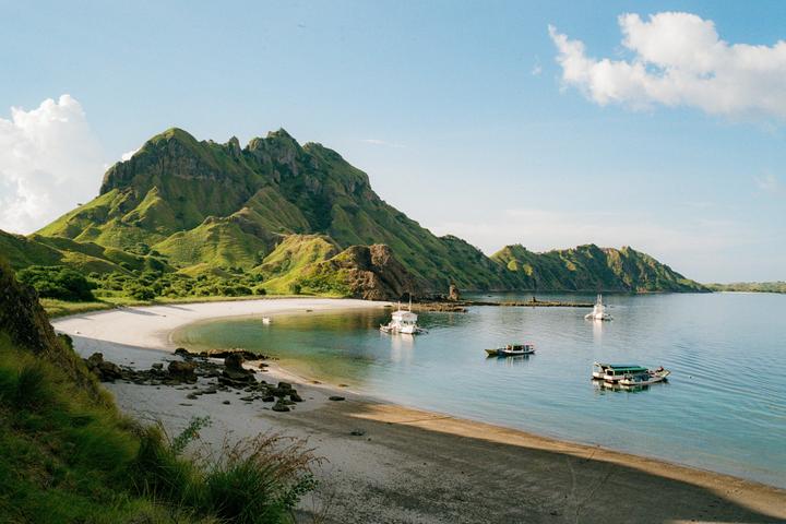 Several boats are anchored in a bay with a white sand beach and green, hilly islands under a blue sky.