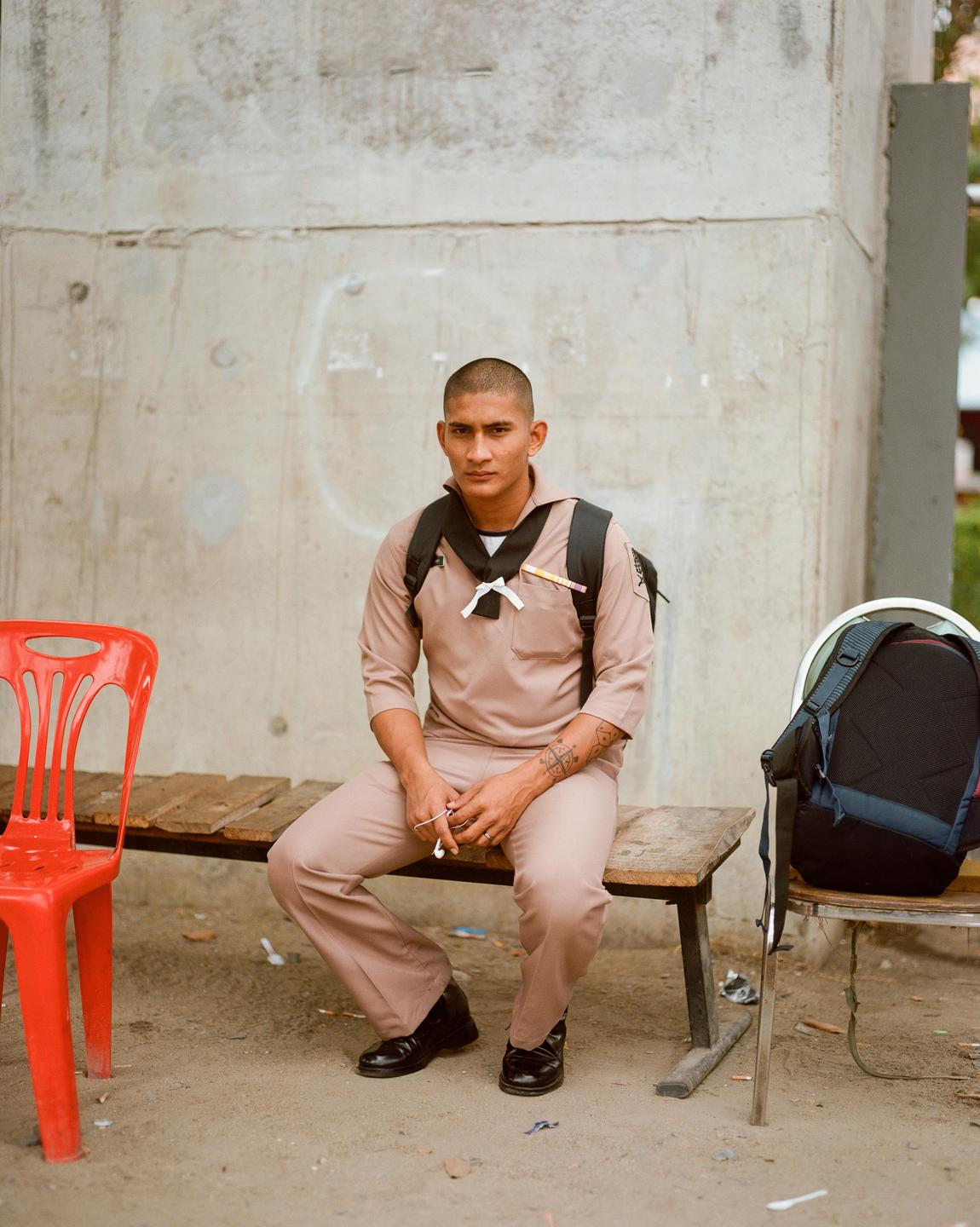 A man in a uniform and backpack sits on a wooden bench next to a red chair.