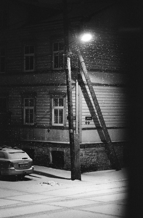 A street light illuminates falling snow next to a building and a parked car.