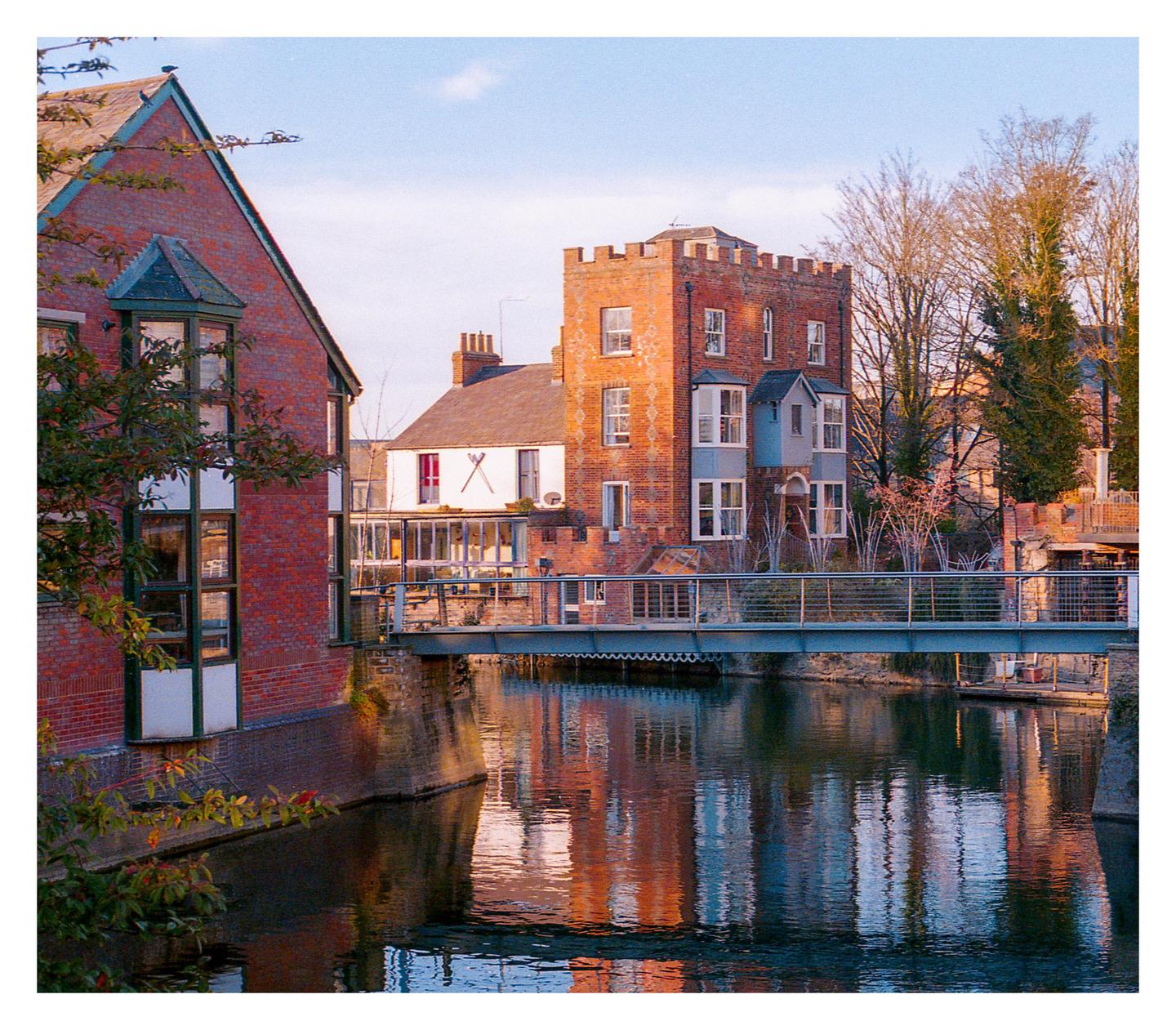 A brick building with a turret stands beside a river with a pedestrian bridge crossing it.