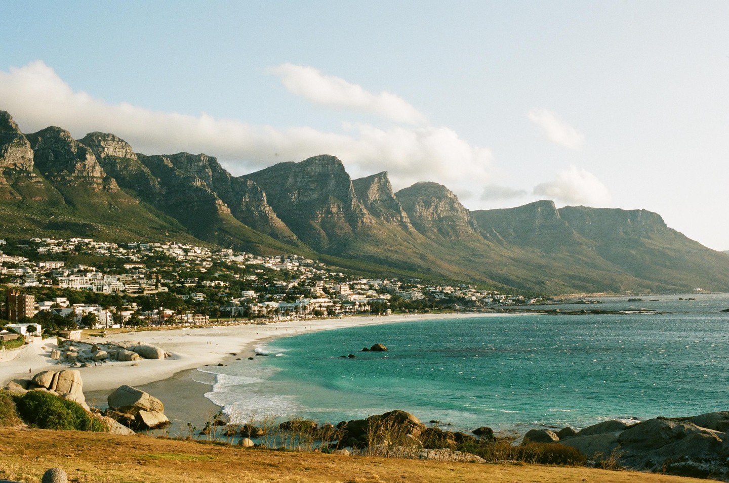 A coastal town with white buildings is nestled at the base of a mountain range, with a sandy beach and turquoise ocean in the foreground.