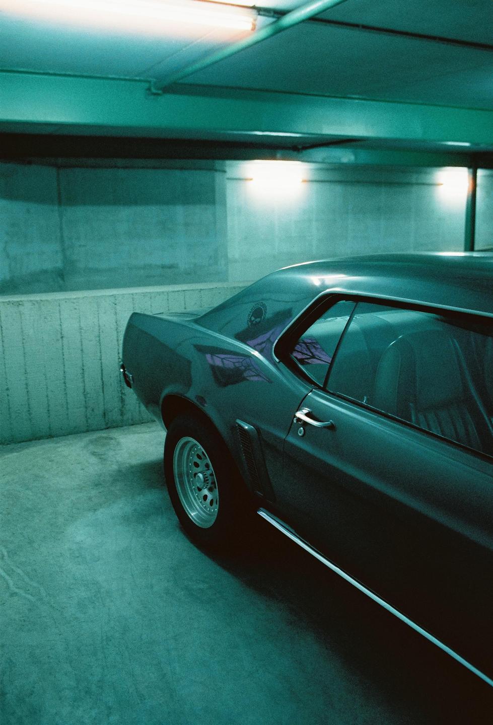 A dark car is parked in a concrete garage with teal lighting.