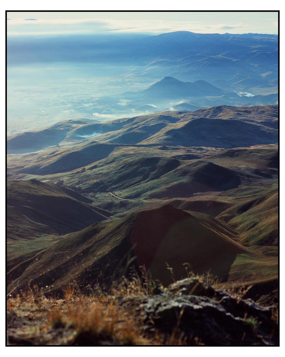 A vast landscape of rolling hills and mountains is seen from a high vantage point, with a hazy sky above.