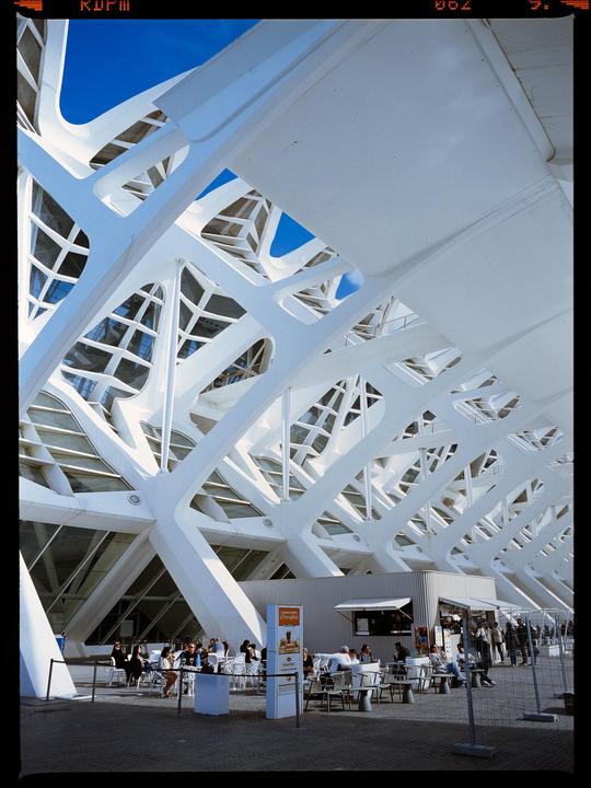 A modern white architectural structure with geometric patterns is visible against a blue sky, with an outdoor cafe and people seated below.