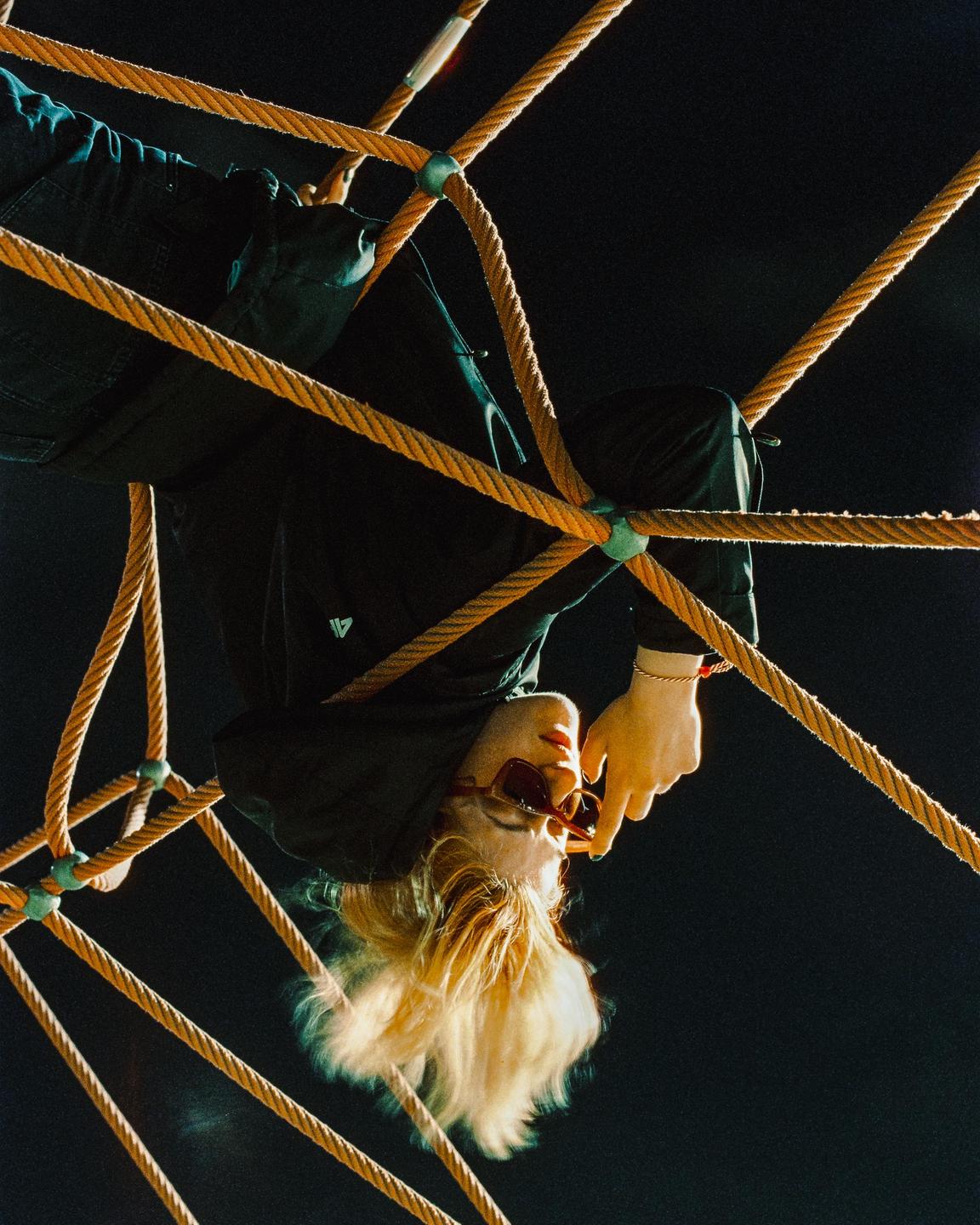 A woman with blonde hair and sunglasses hangs upside down in a rope climbing net at night.