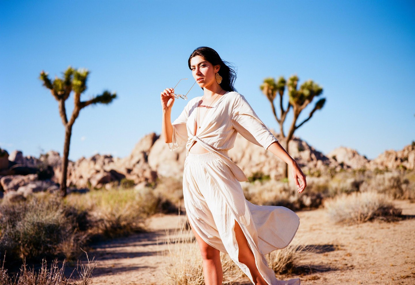 A woman in a light-colored outfit holds sunglasses in a desert landscape with Joshua trees and rocks.