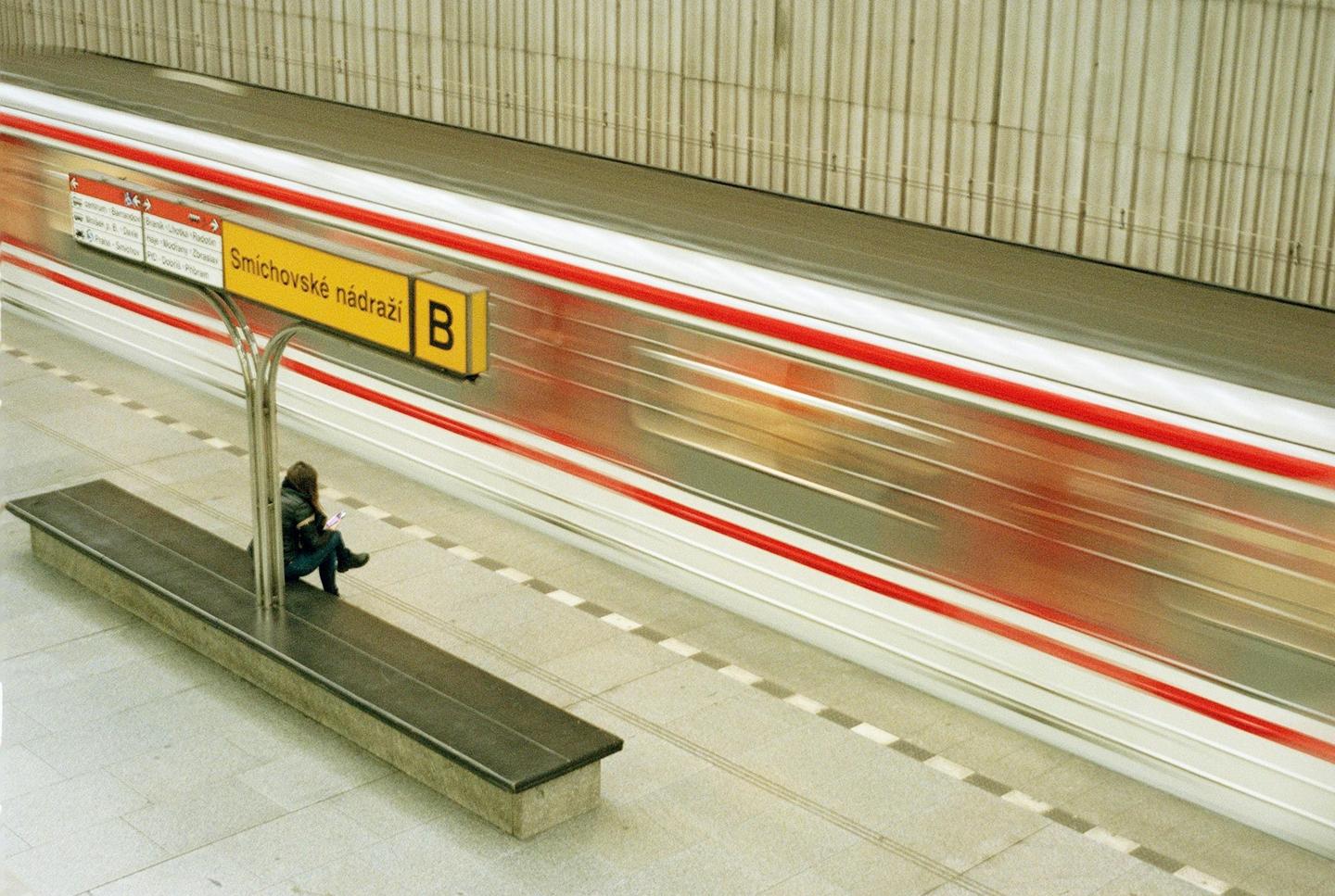 A woman sits on a bench at a subway station as a train speeds by, creating a motion blur effect.