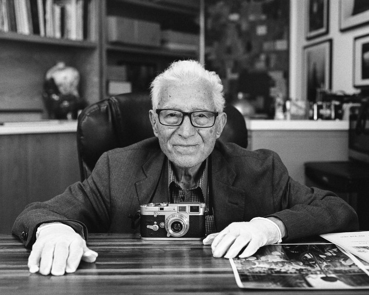 An elderly man wearing glasses and white gloves sits at a desk with a camera in front of him, smiling at the viewer.
