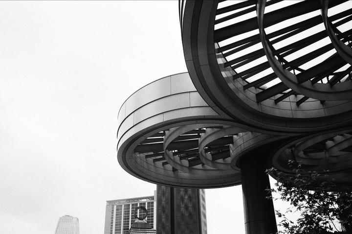 A low-angle view shows the circular architectural details of a modern building against a bright sky, with other buildings and foliage visible in the background.