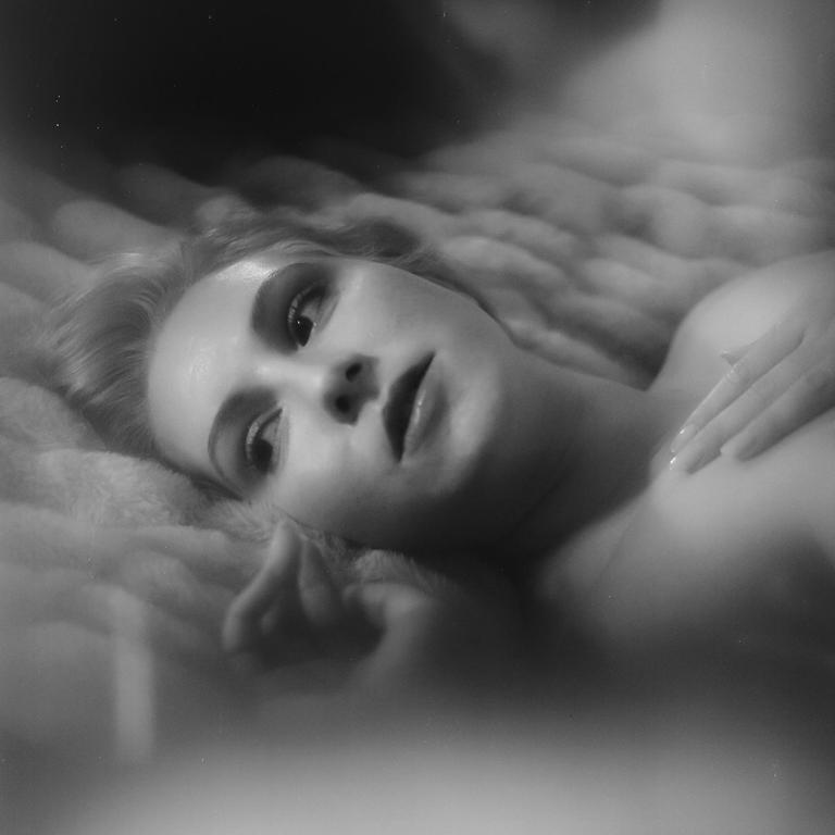 A woman with light hair lies on a textured surface, looking upwards with one hand resting on her chest.
