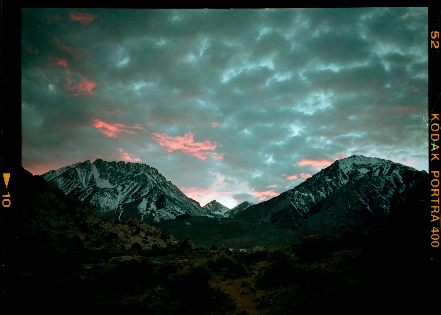 A dramatic sunset with pink clouds illuminates snow-capped mountains and a dark valley.