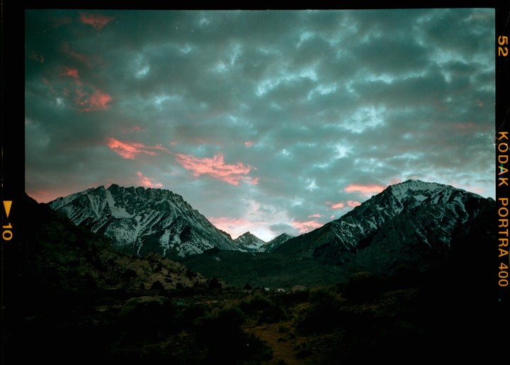 A dramatic sunset with pink clouds illuminates snow-capped mountains and a dark valley.