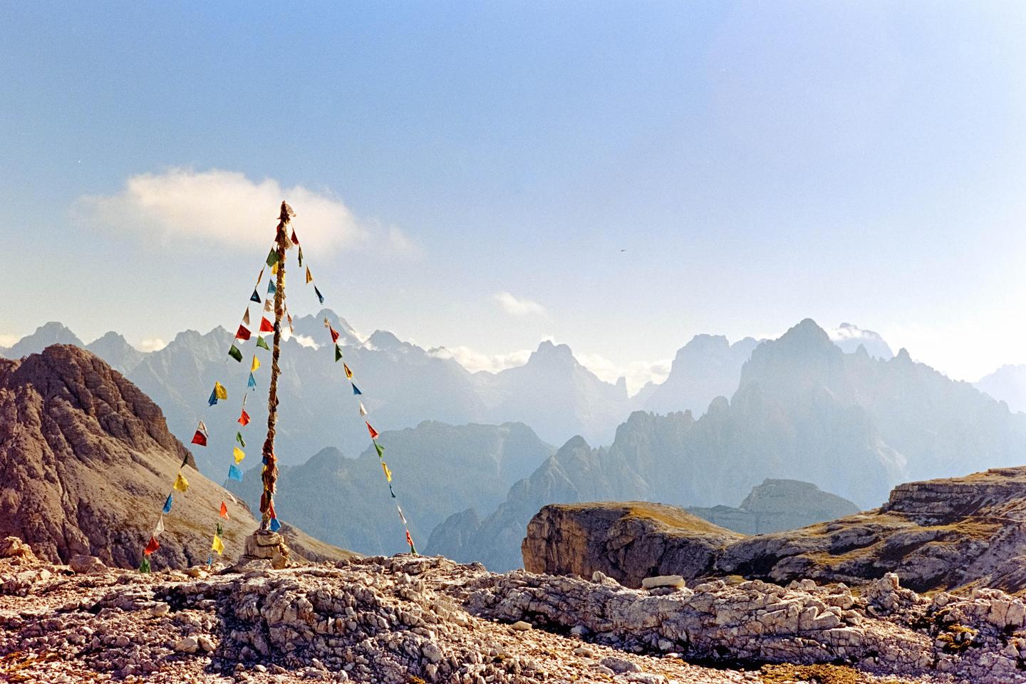 A pole adorned with colorful prayer flags stands on a rocky mountain peak, with a range of mountains in the hazy distance under a clear sky.
