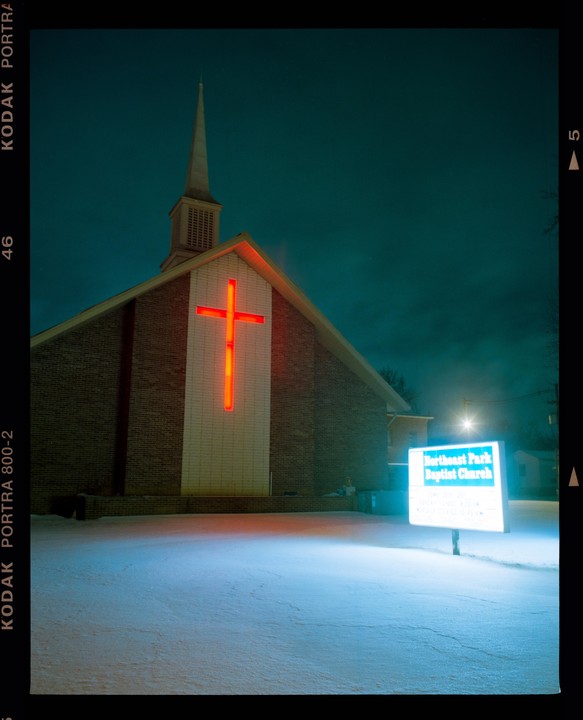 A church with a glowing red cross and a lit sign stands in a snowy landscape at night.