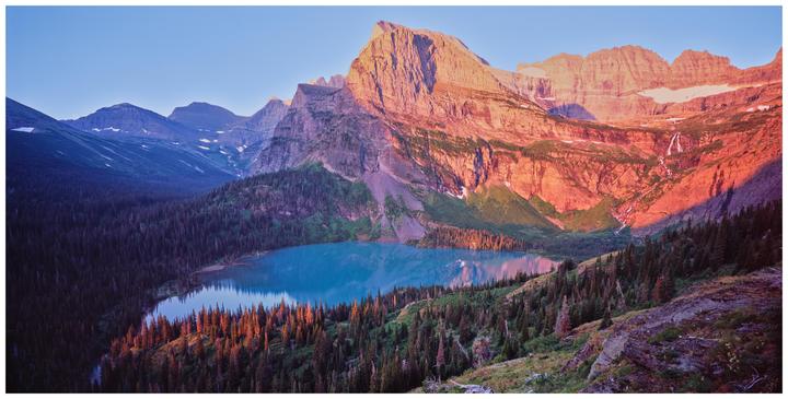 A mountain landscape shows a lake surrounded by pine trees with a waterfall cascading down a distant mountain.