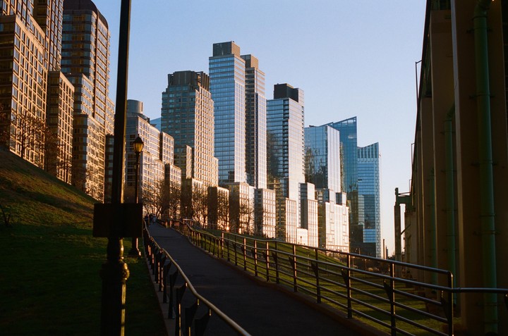 A paved path with a metal railing runs alongside a grassy hill, with tall buildings and a large overpass visible in the background.